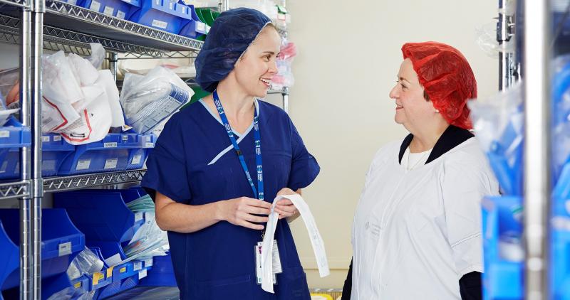 Two women speak in a hospital supply room.
